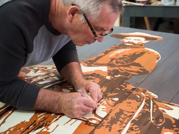 man carefully attends to a detail of the completed stencil painted cowboy with a precision knife