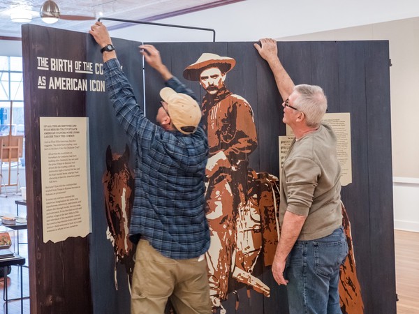 two people hold up and place metal brace across two large wooden panels of cowboy image