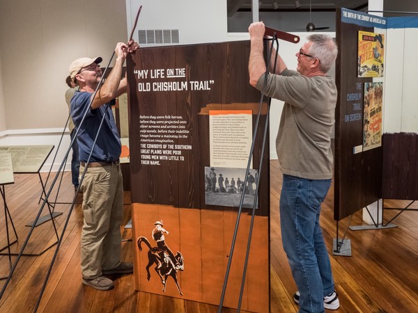 two people attach leather straps to hang wooden panel from metal frame; stenciled image of rider on bucking bronco over orange background on panel