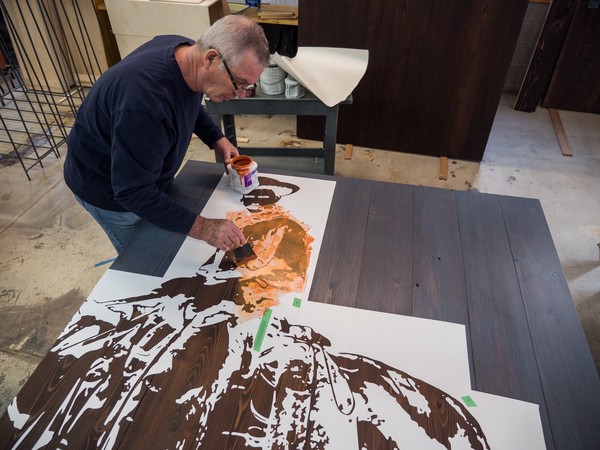 man paints orange stencil layer of cowboy image on dark stained cedar panel