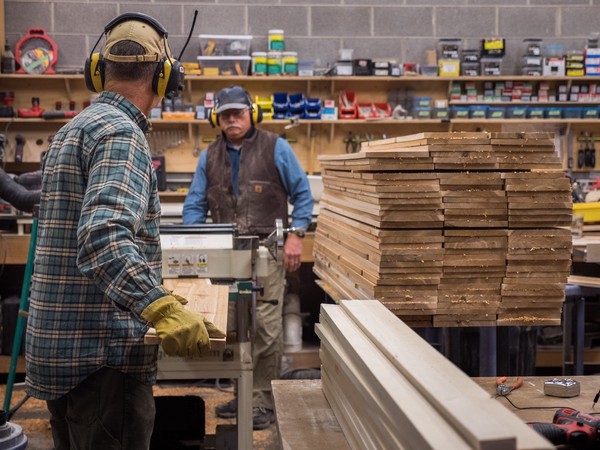 two carpenters feed a cedar board through a planer next to a large stack of boards