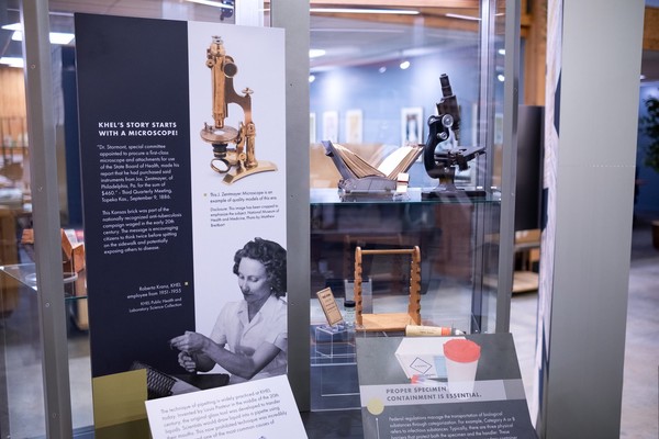 A microscope in the case alongside the origin story of KDHE on a printed panel.