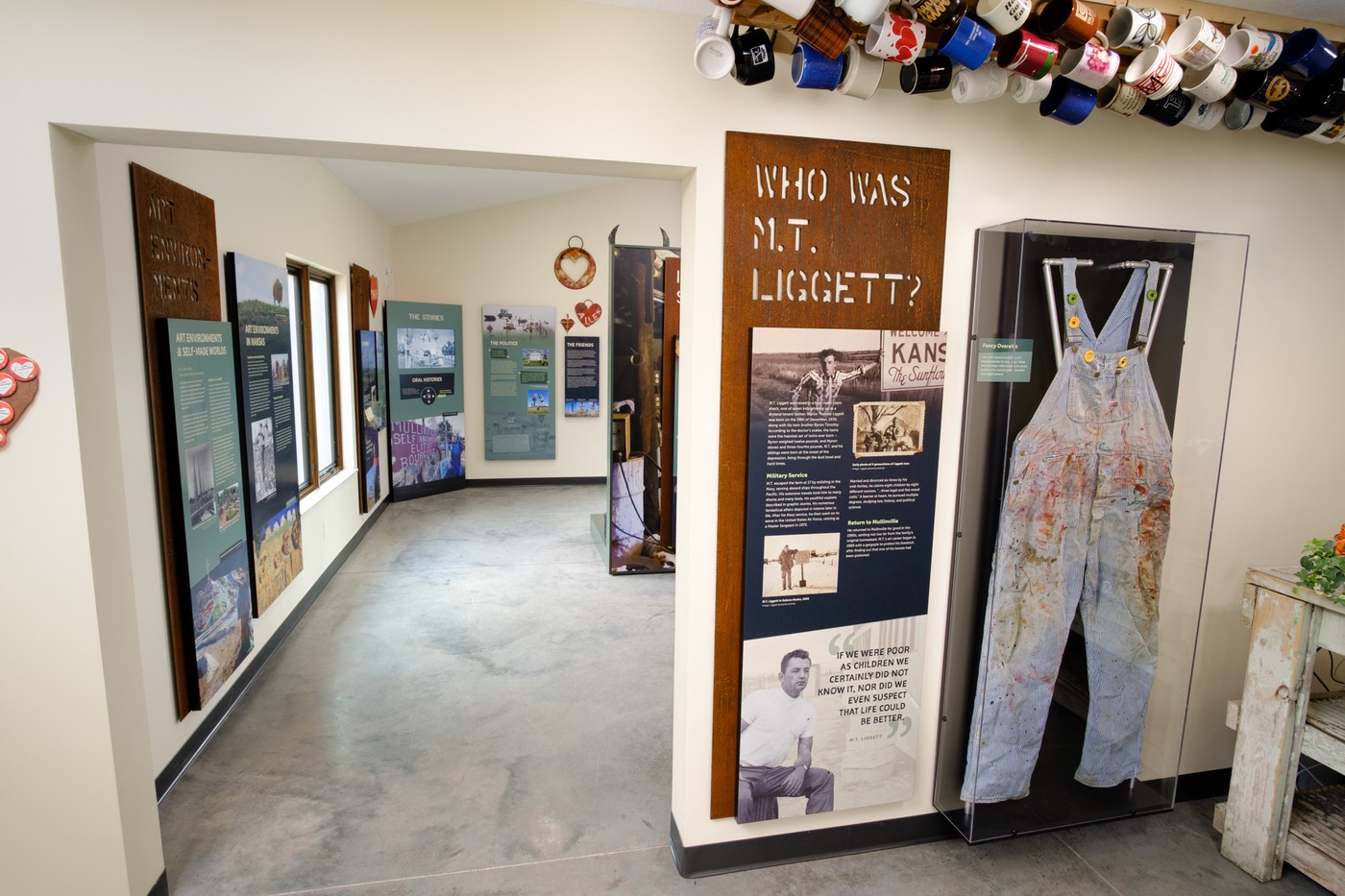 Rusted metal exhibit panel titled 'Who was M.T. Liggett' next to a paint-stained pair of overalls displayed within a case