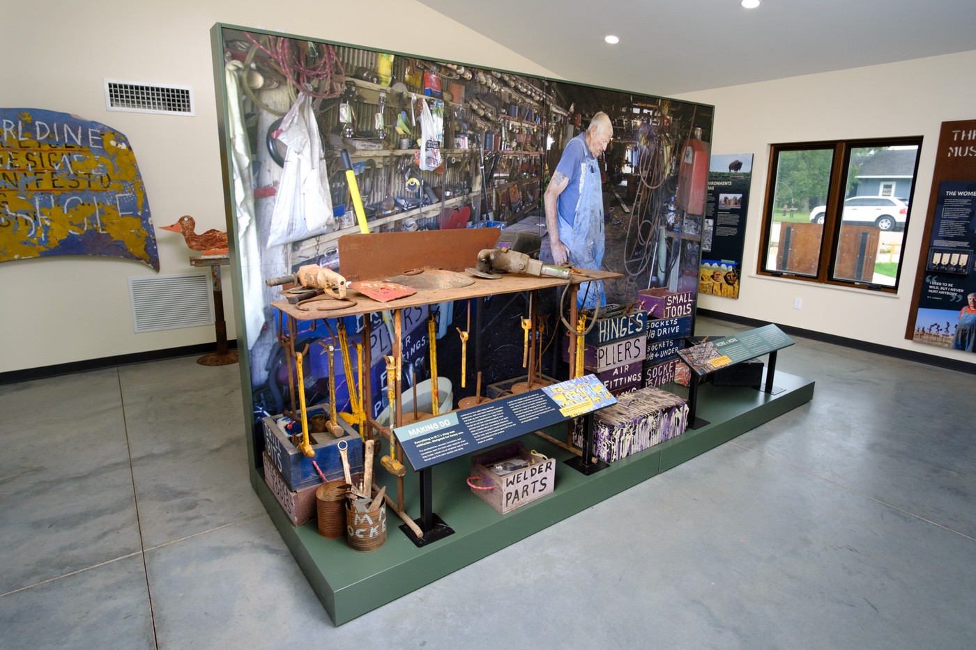 Second side of center island shows another large photo of Liggett's studio with him in it. In front of the photo are tools, a metal workbench, and storage crates painted blue and purple and hand labelled.