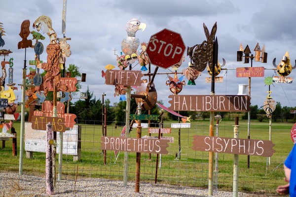 Welded, painted, and weathered metal sculptures mounted on poles along a fence line; one artwork features old stop signs