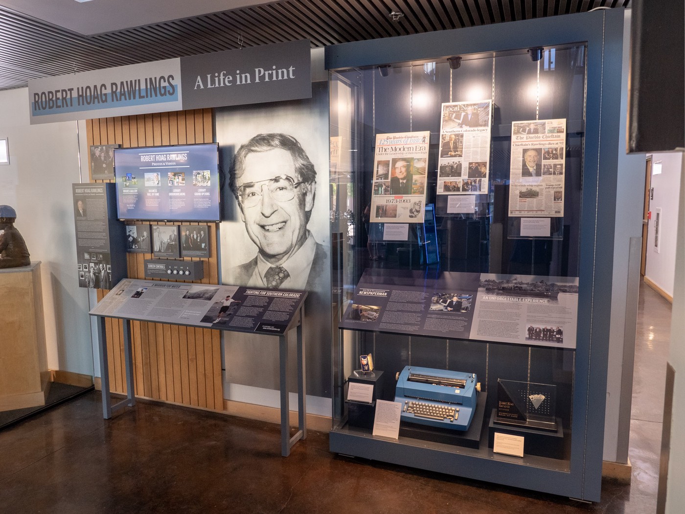 exhibit filling a wall featuring steely blue colors; photo and interpretive panels, screen, large metalic portrait, display case
